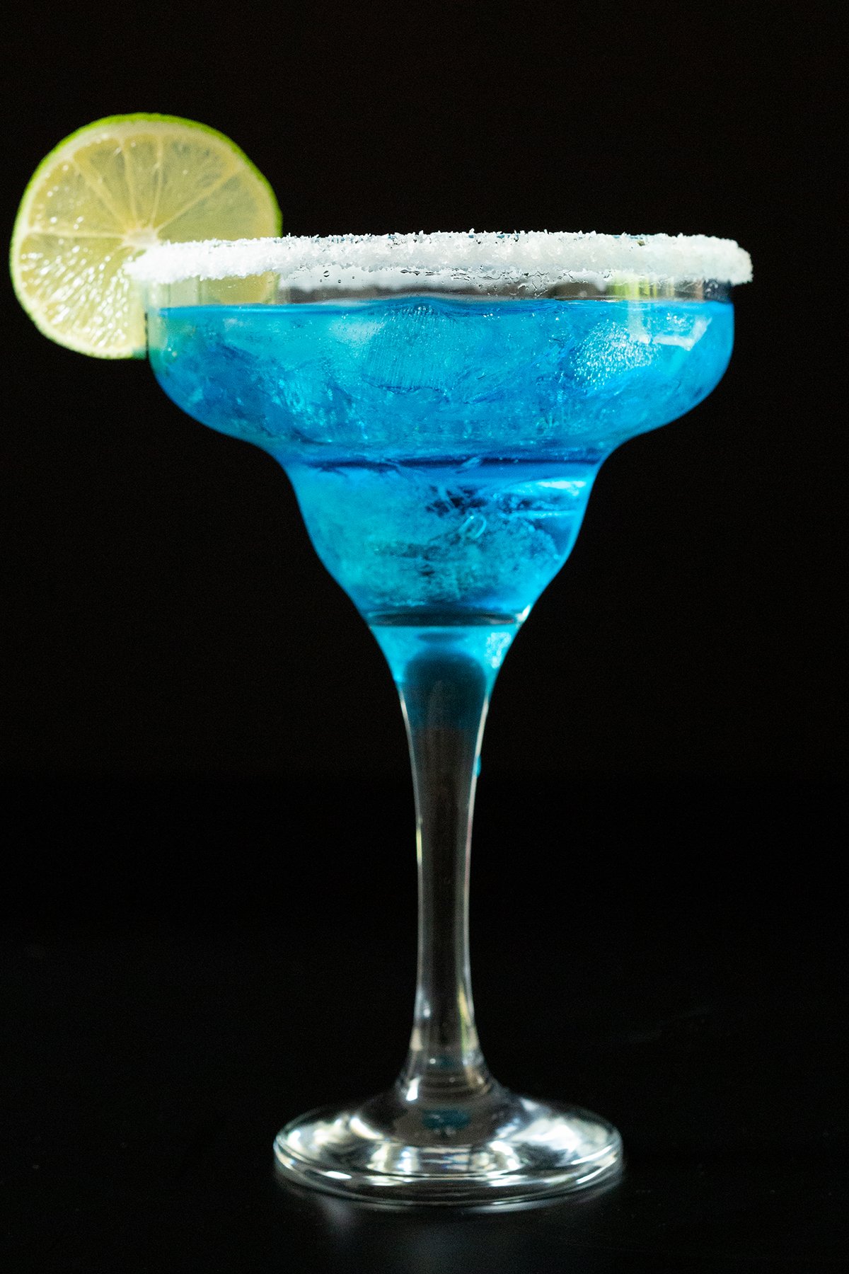 A tall margarita glass is filled with a vibrant bright blue margarita. It's garnished with a salt rim and a lime wheel.