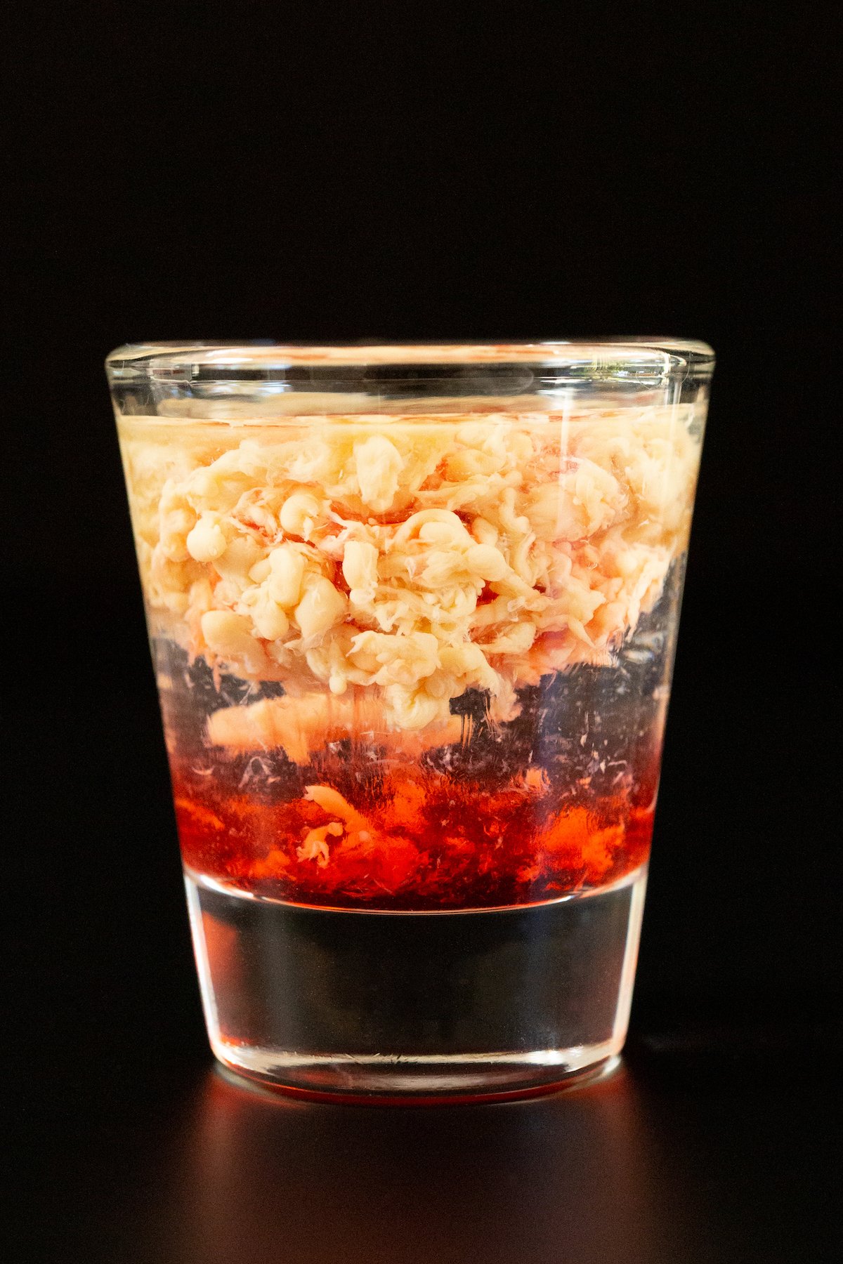 A single shot glass filled with a layered shot that's red on the bottom, clear in the middle, and the top is brown and textured like a brain.