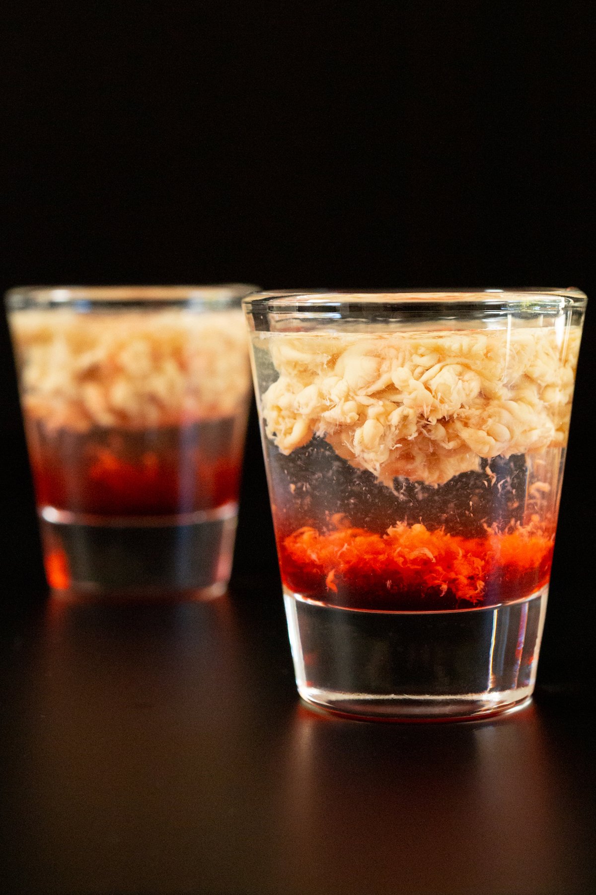 Two shot glasses, on is in focus in the foreground and the other out of focus in the background. They are filled with a bottom red layer, a clear middle layer, and a top brown layer that looks like brains.