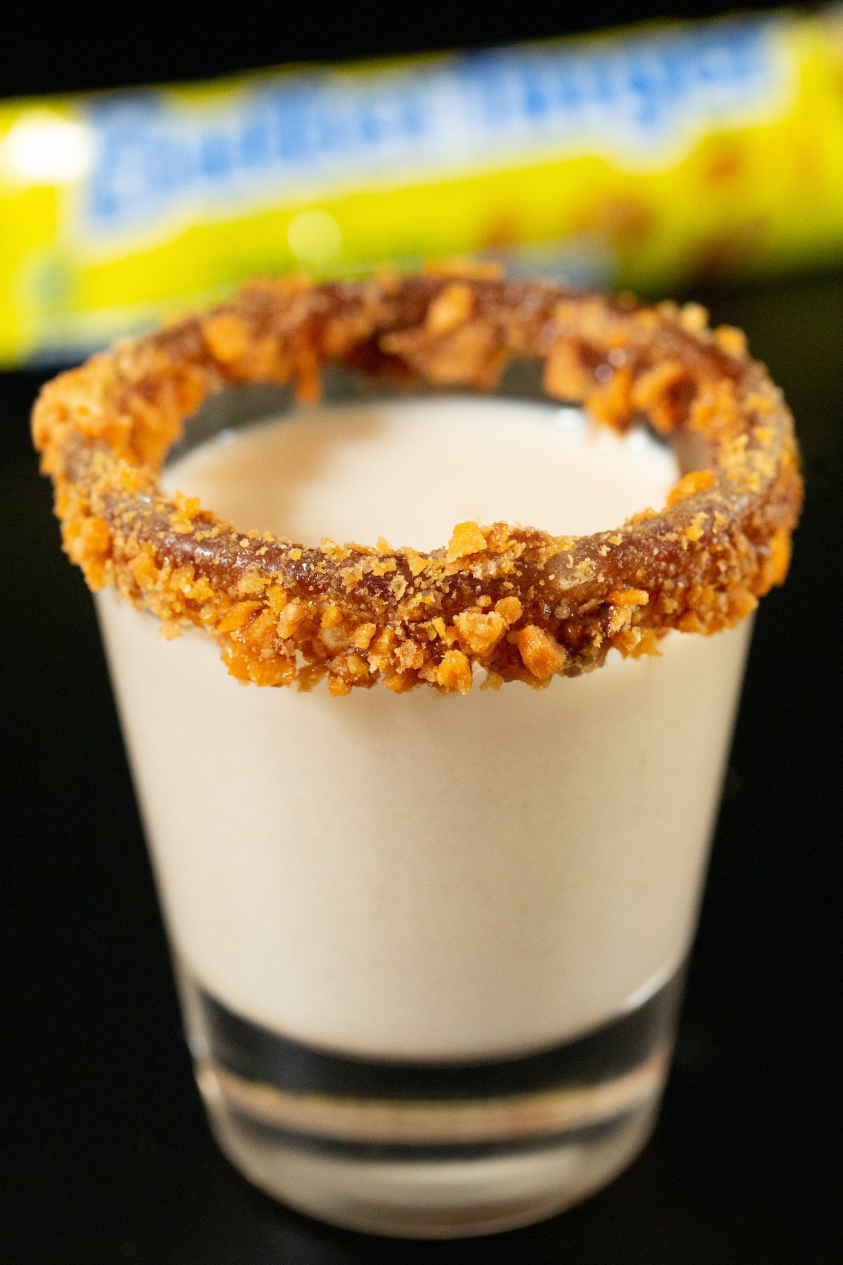 A shot glass is filled with a light brown Butterfinger shot. The rim is coated in chocolate sauce and crushed candy bar. 