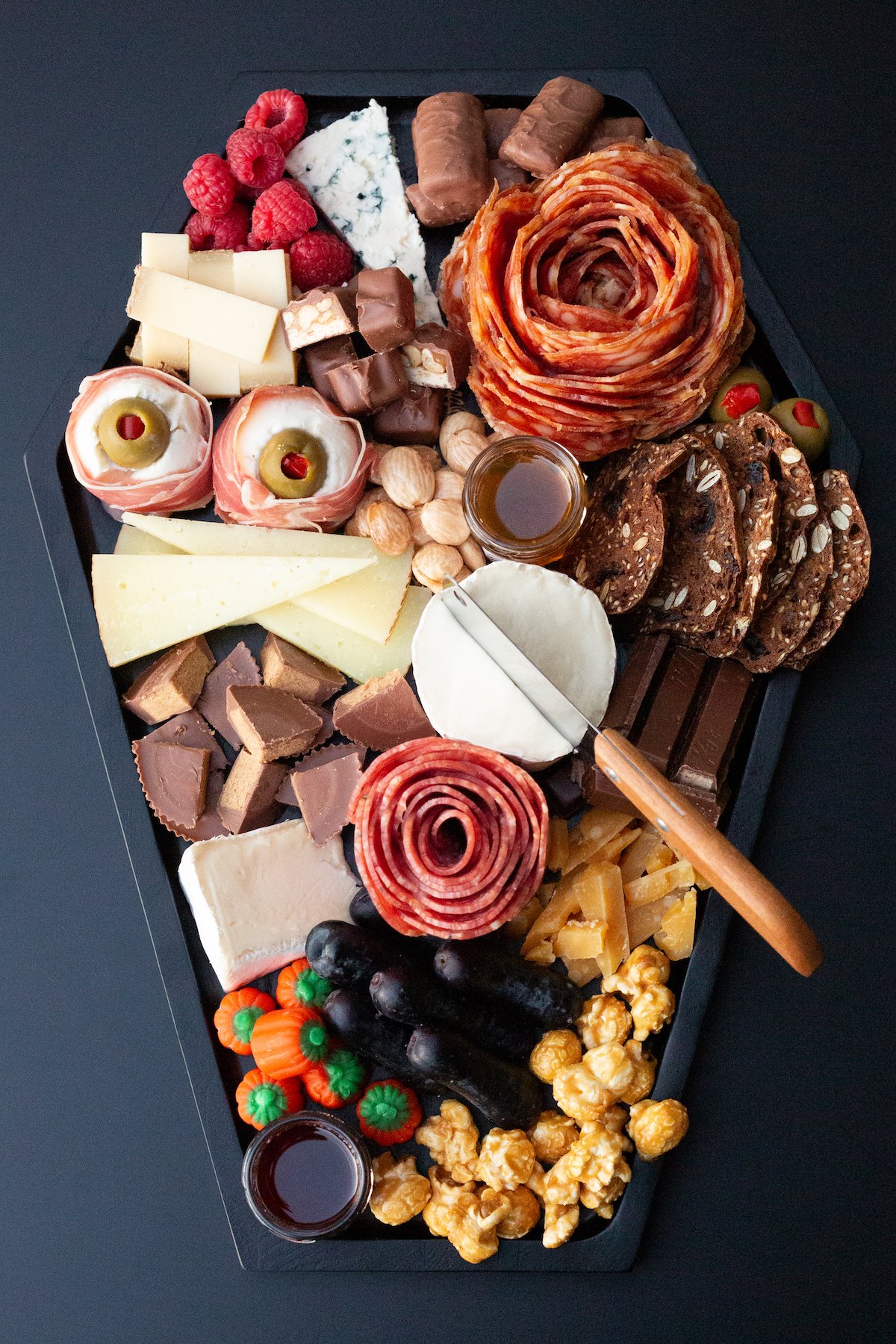 A black, coffin shaped cheese board is filled with two meat roses, chopped various Halloween candies, small jars of jam & honey, several cheeses, grapes, and nuts.