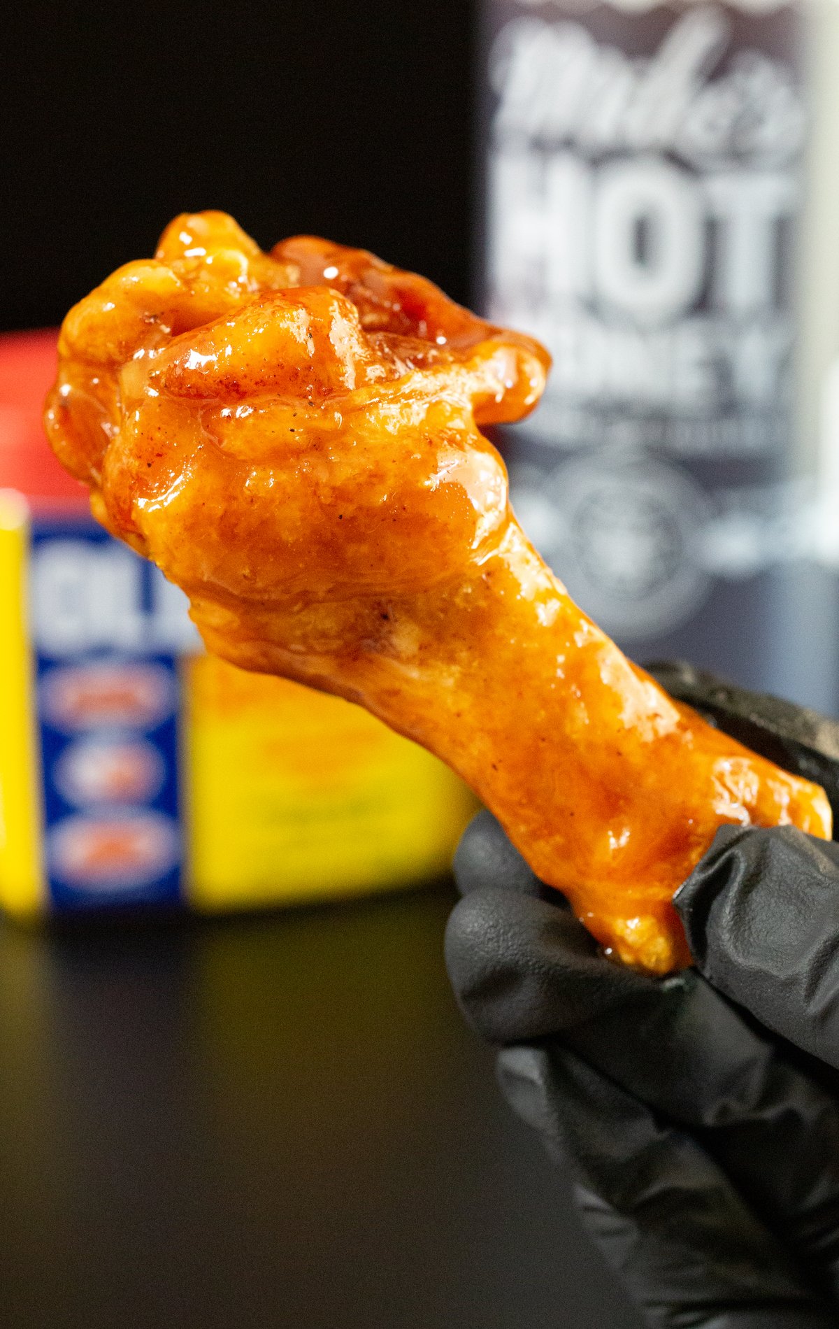 A hand olds up a drumstick chicken wing covered in a shiny glaze. In the background, a bottle of Mike's Hot Honey and and Old Bay seasoning are out of focus.