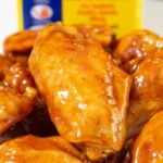 Close up of a flat chicken wing covered in a reddish-orange glaze. An Old Bay seasoning container is out of focus in the background.