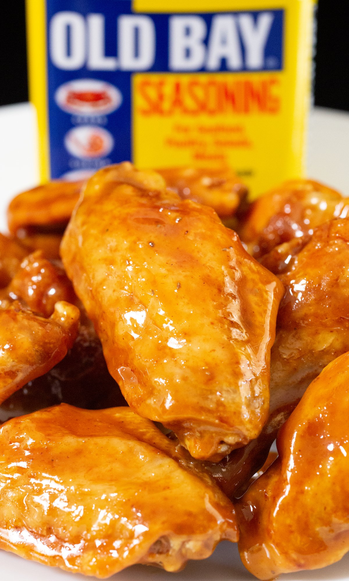 Close up of a flat chicken wing covered in a reddish-orange glaze. An Old Bay seasoning container is out of focus in the background.