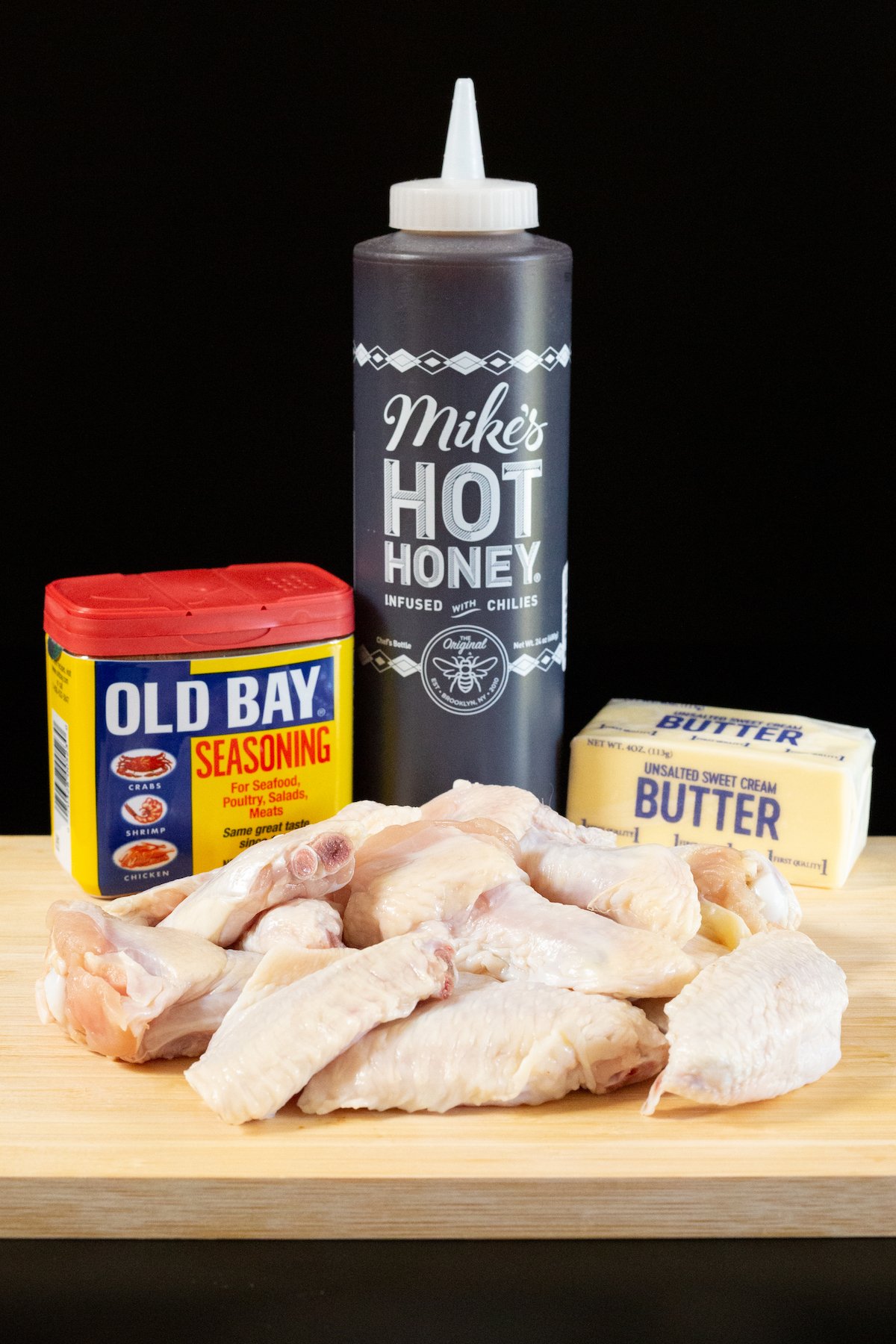 A bottle of Old Bay seasoning, Mike's Hot Honey, a stick of butter, and about a dozen chicken wings on a wood cutting board.
