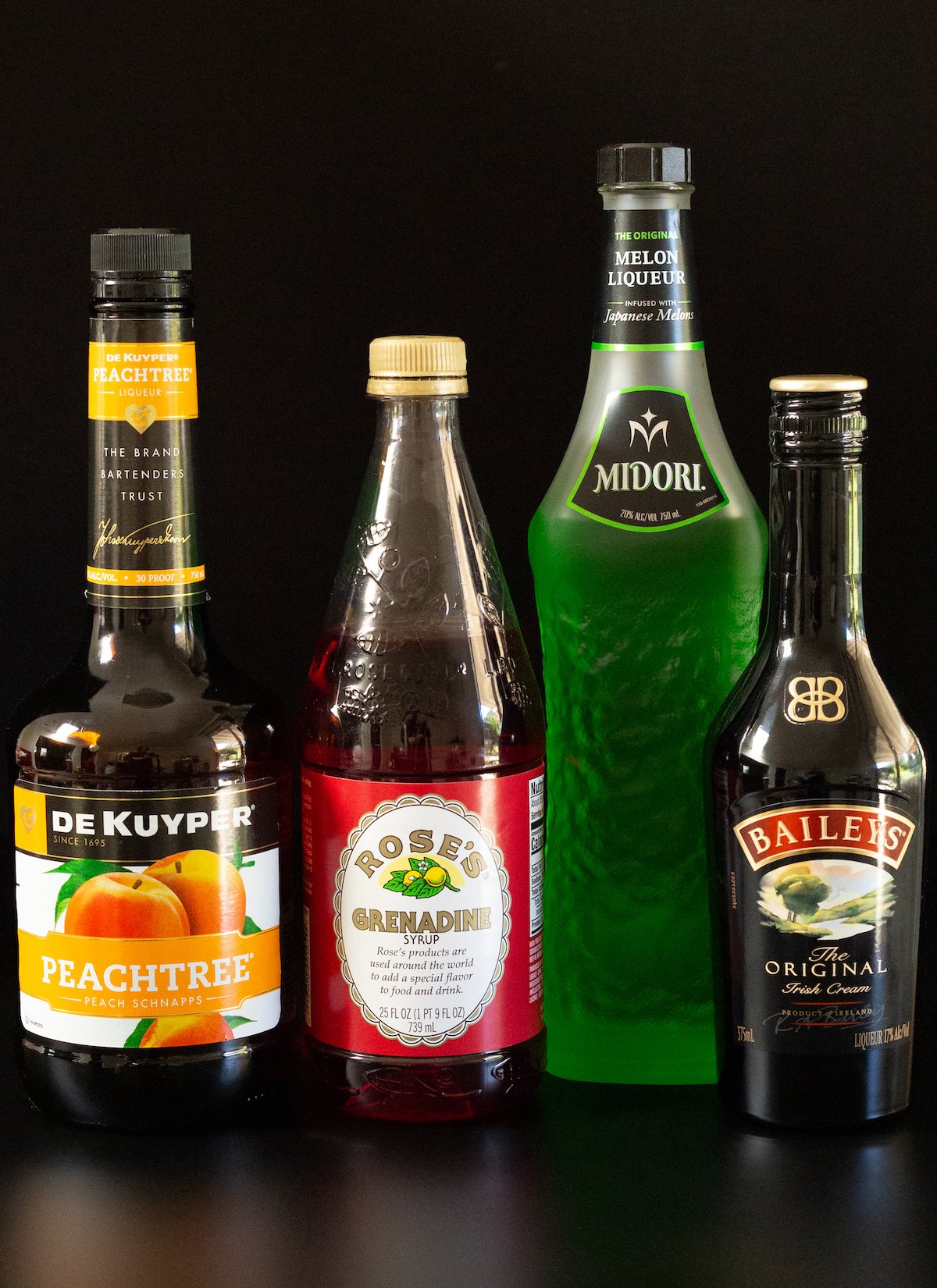 A bottle of peach schnapps, Rose's grenadine, Midori, and Bailey's Irish Cream on a black background.