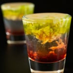 Two shot glasses with the bottom filled with a red liquid, middle is clear, and top is brown and green with the texture of brains.