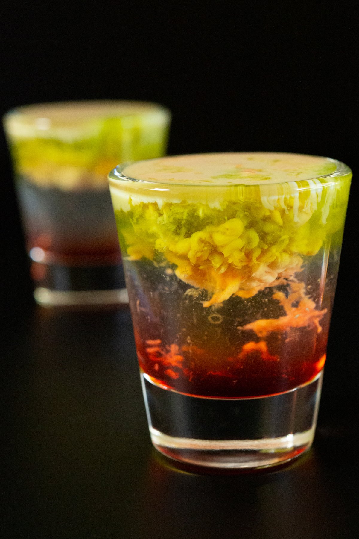 Two shot glasses with the bottom filled with a red liquid, middle is clear, and top is brown and green with the texture of brains.