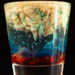 A shot glass is filled with a layered shot that is red on the bottom, then blue, then clear, and a light brown brain texture for the top layer.