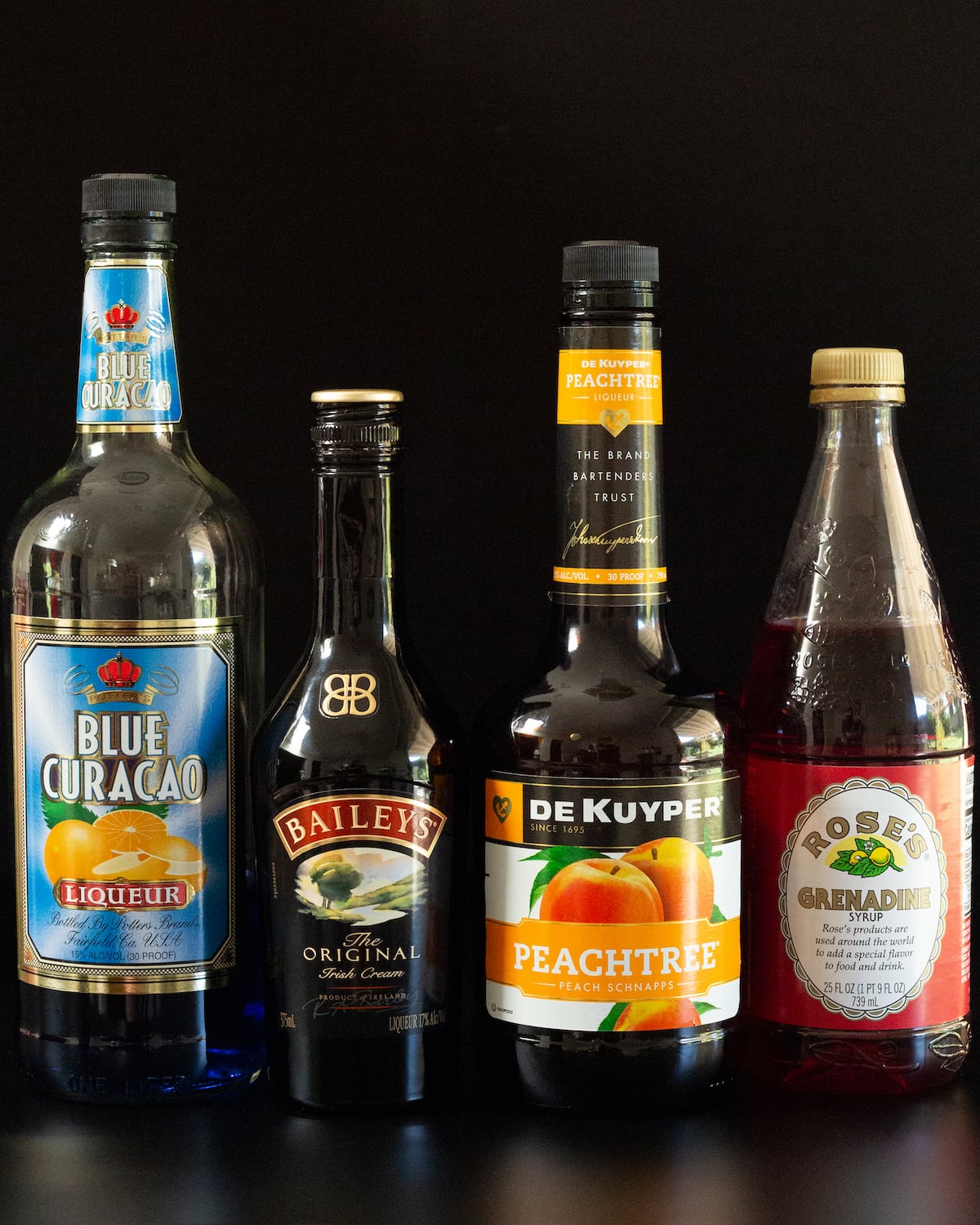 A bottle of blue curacao, Bailey's Irish Cream, Peach Schnapps, and grenadine all on a black background.