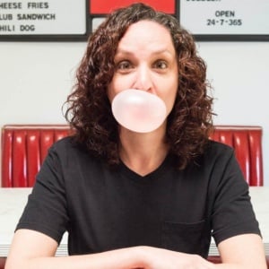 A women in a black t-shirt sits at a red diner chair and blows a bubble of pink bubblegum.