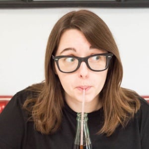 A woman in a black t-shirt sips on a coke in a bottle while sitting at a diner table.