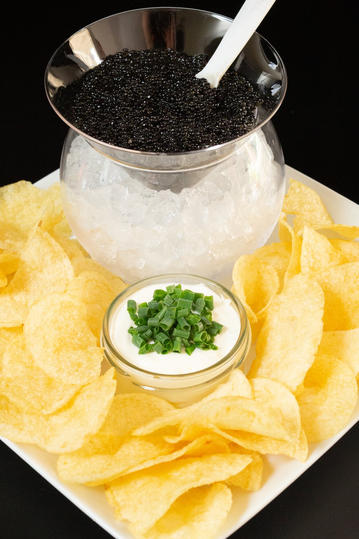 A circle glass orb is filled with pebble ice and a round insert with a triangle bottom is filled with caviar and inserted on top. It's on a white platter filled with potato chips and a small glass jar with creme fraiche and sliced green onions.