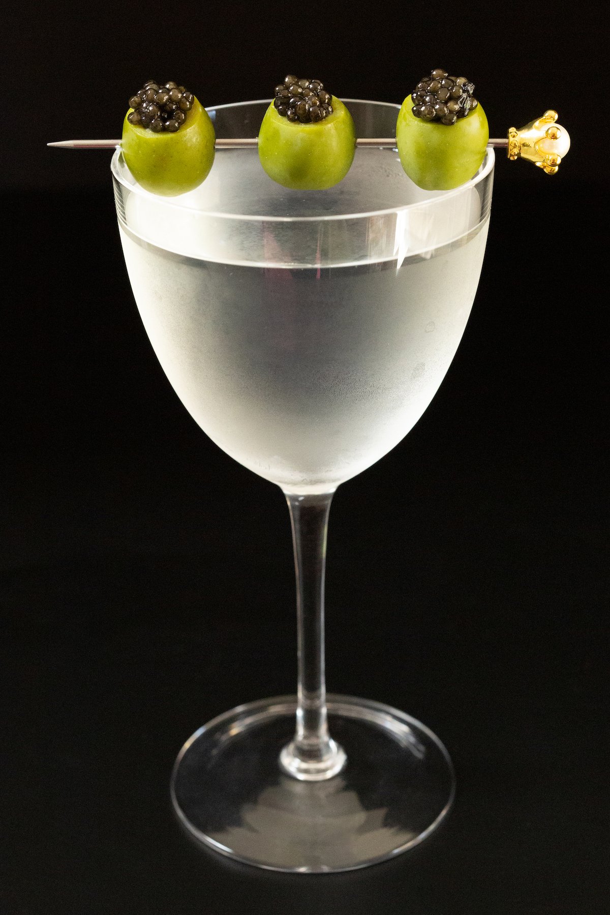 A frosted Nick & Nora glass filled with a martini that's garnished with a cocktail pick with three skewered green olives stuffed with caviar.