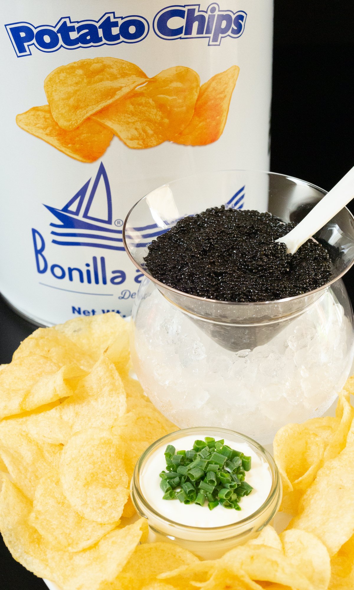 A tin of potato chips, a glass caviar chiller with ice on the bottom and caviar on top, and a plate of potato chips with a jar of creme fraiche topped with sliced green onions.