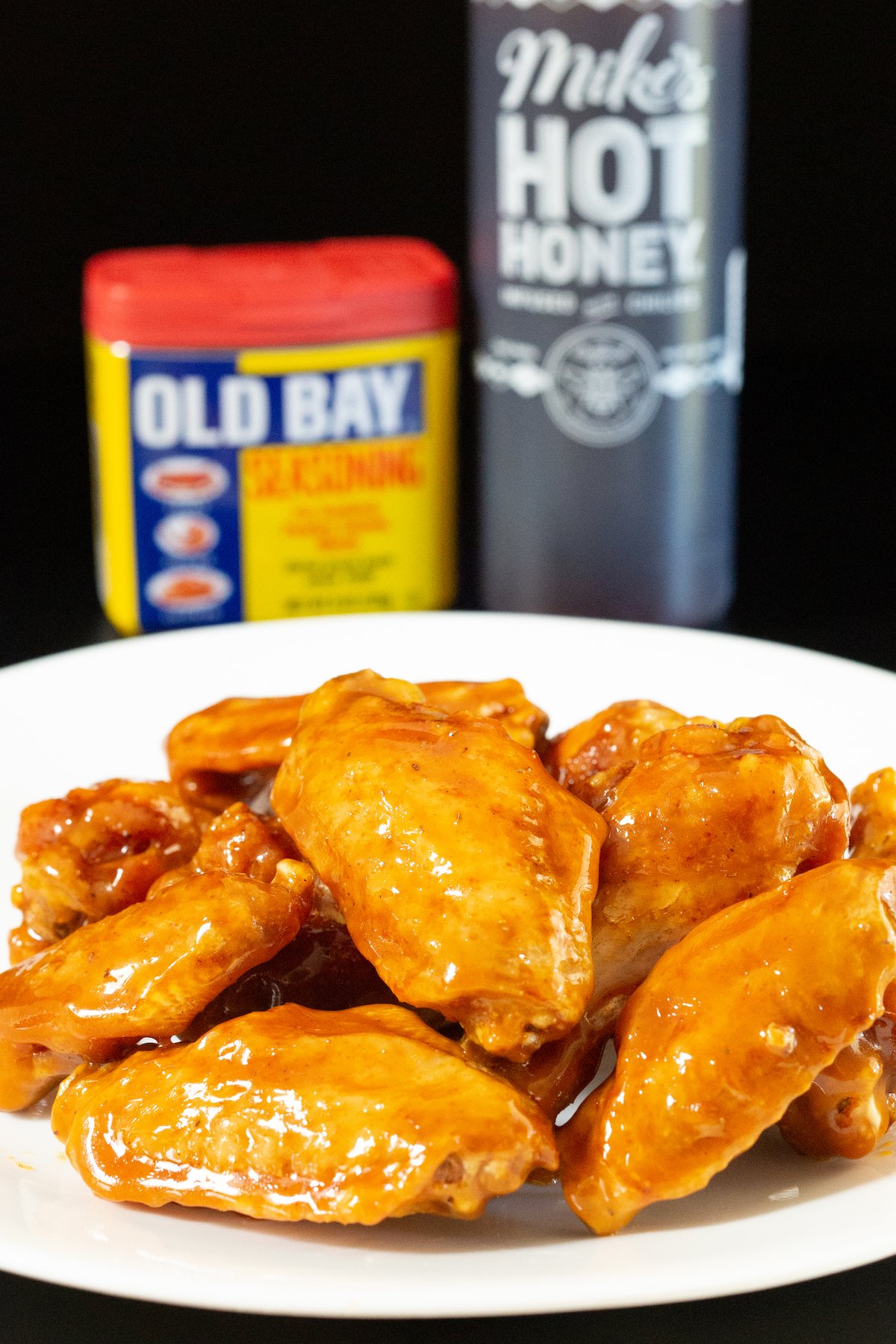 A dinner plate filled with about a dozen chicken wings covered in an orange colored glaze. An old bay seasoning container and a bottle of hot honey are out of focus in the background.