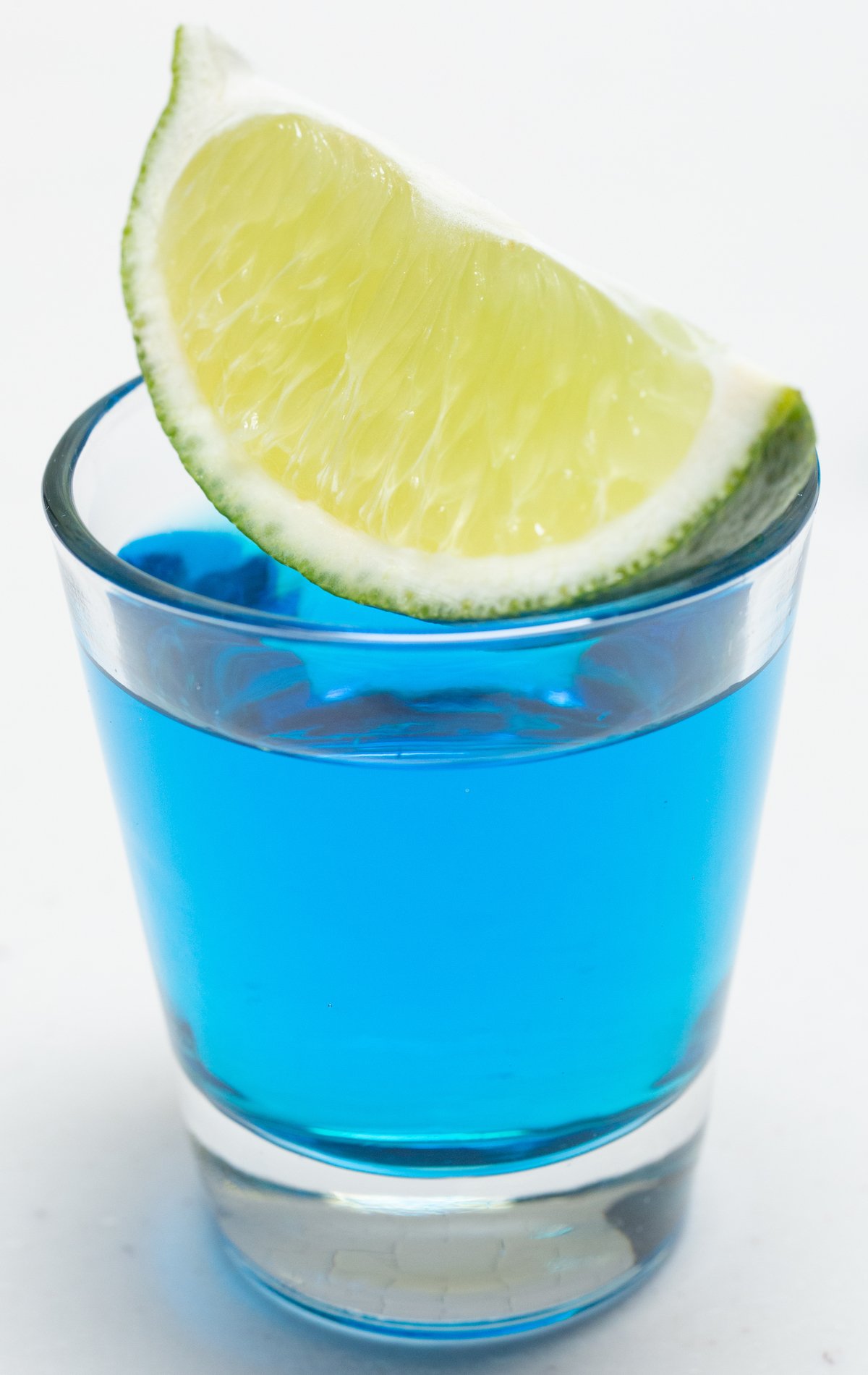 A shot glass filled with a bright blue shot has a lime wedge sitting across the top.