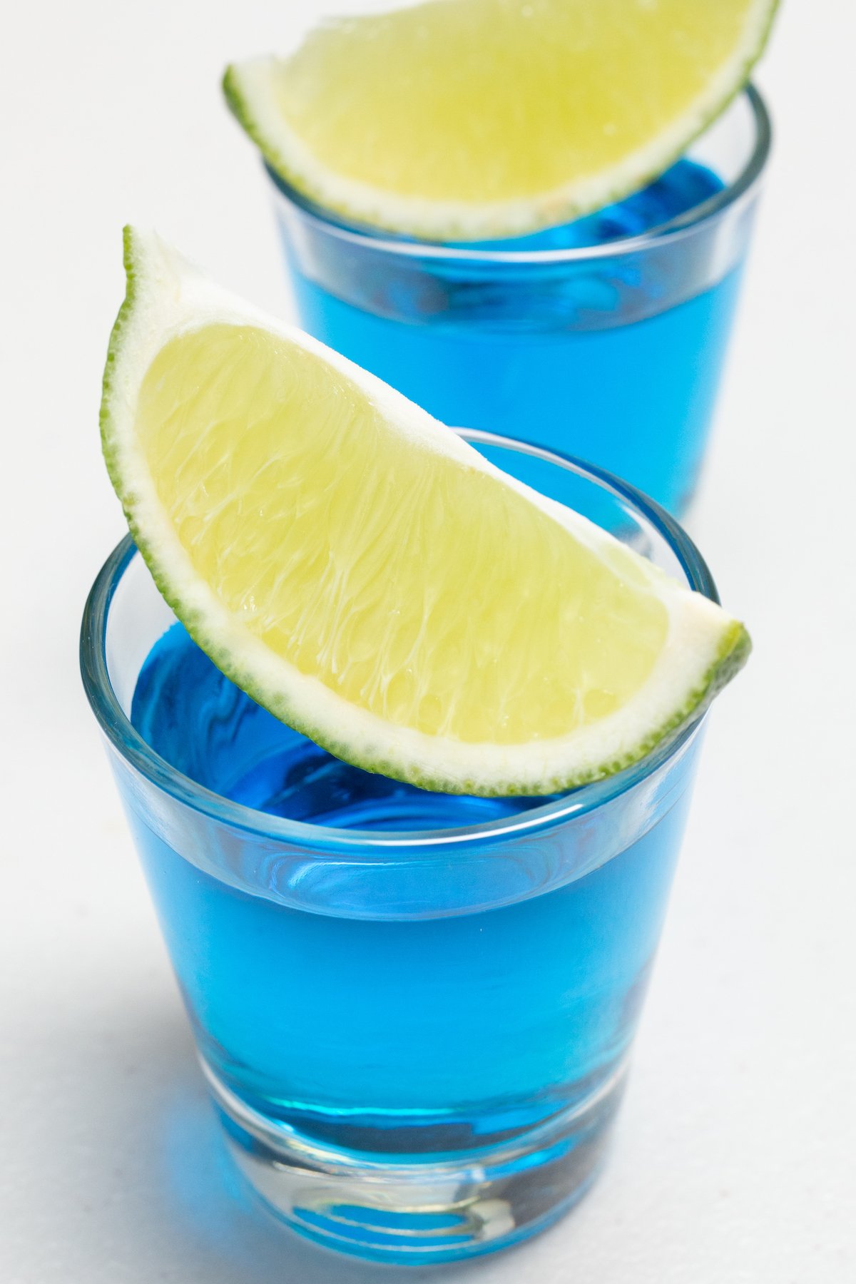 Two shot glasses filled with bright blue liquid are on a white background. Fresh cut lime wedges sit across the top of each shot glass.