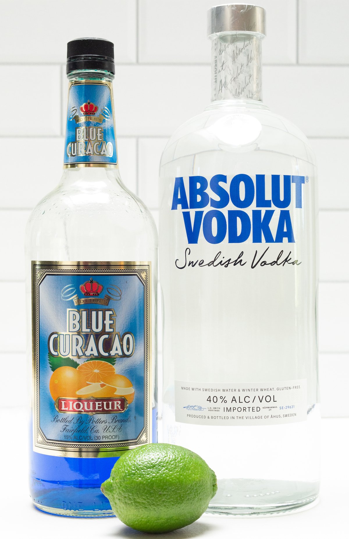 A bottle of blue curacao and a bottle of vodka in front of a white tile background.