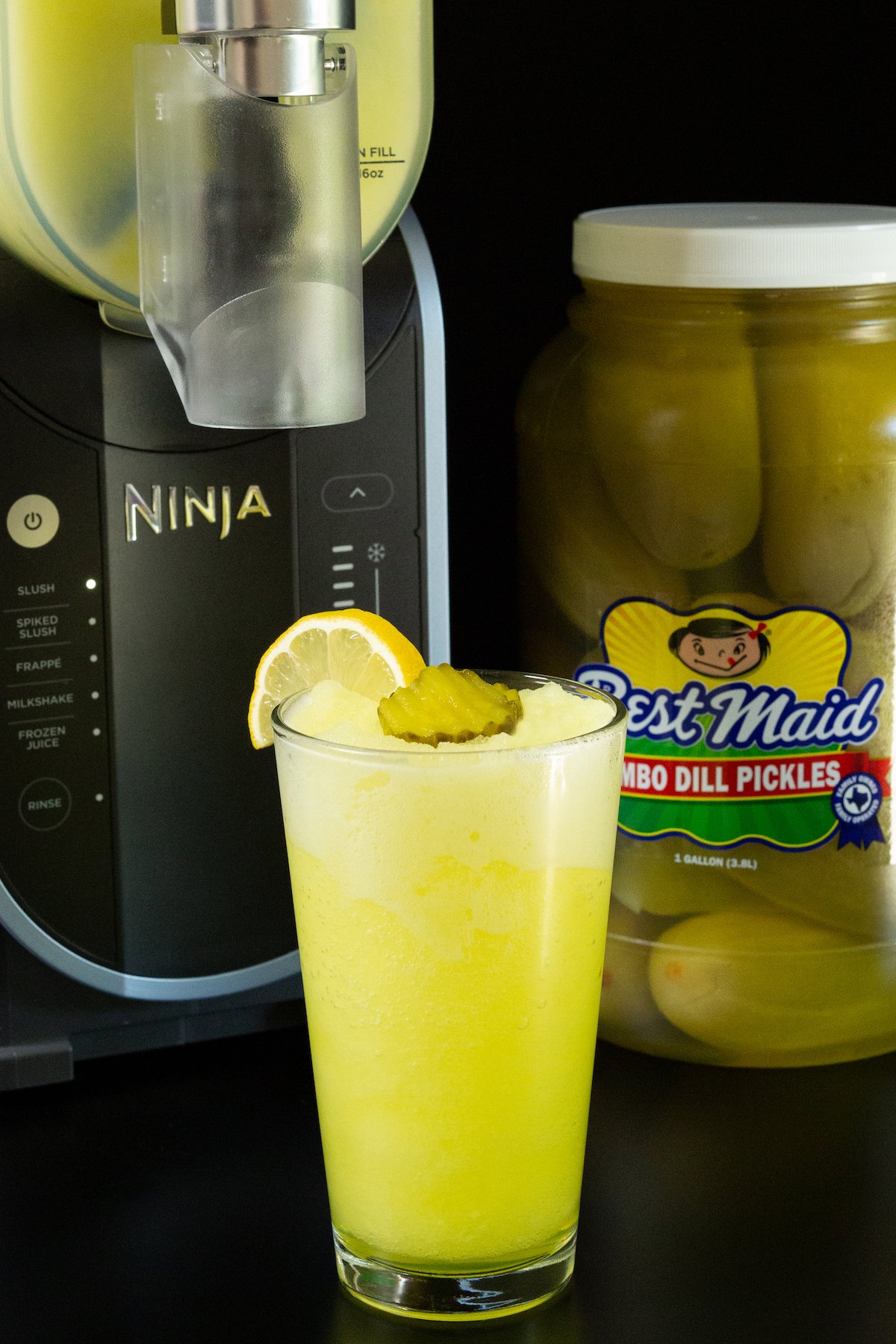 A pint glass fillled with a bright yellow pickle slushi sits in front of a large jar of dill pickles and the Ninja Slushi machine.