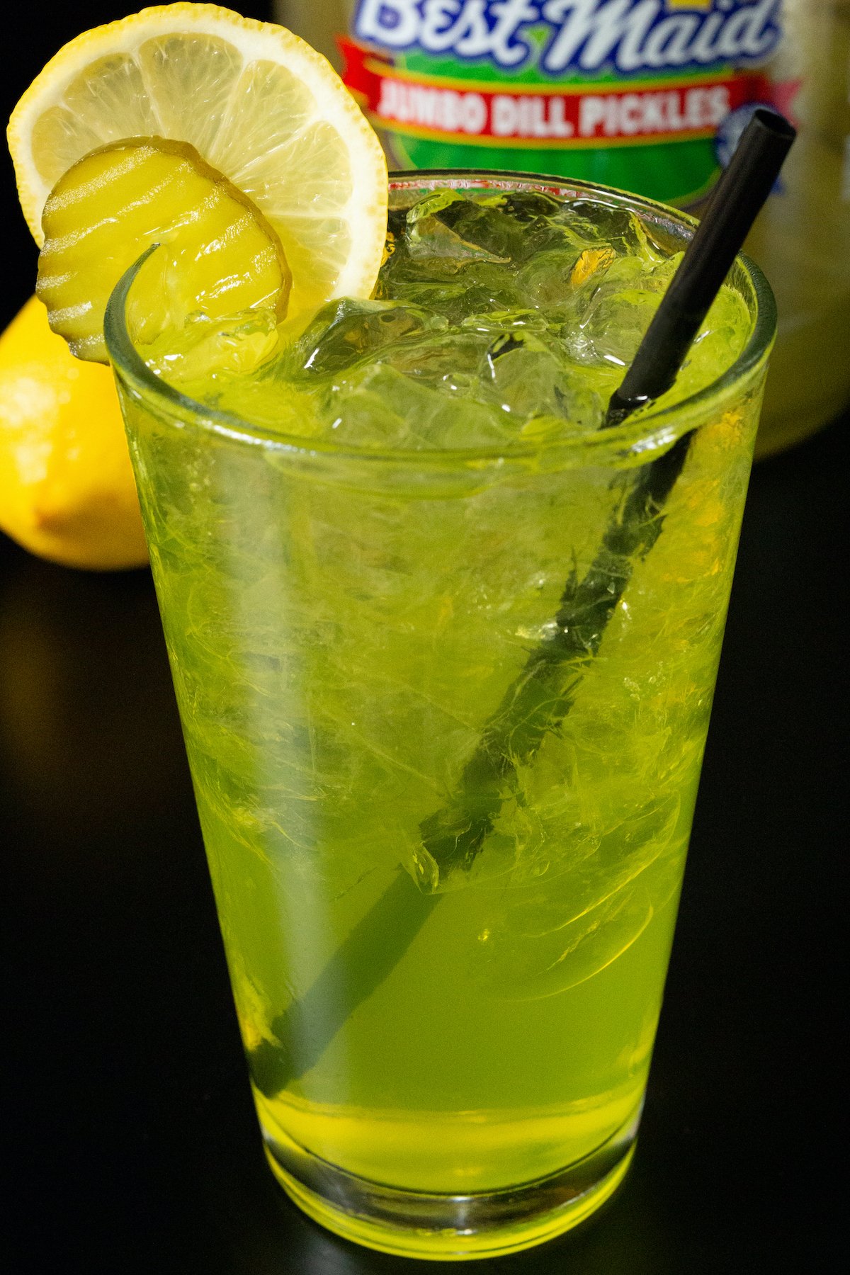 A pint glass filled with clear ice cubes and a bright yellow pickle lemonade drink that's garnished with a pickle chip and a lemon wheel.
