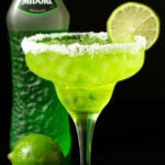 A tall margarita glass is rimmed with coarse salt, filled with a bright green margartia, and garnished with a lime wheel. Out of focus in the background is a bottle of Midori and a whole lime.