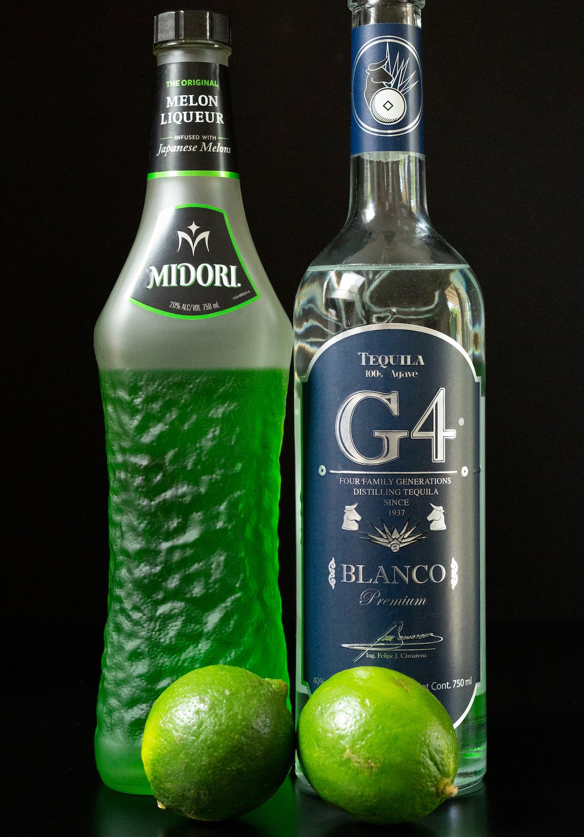 A bottle of Midori and a bottle of G4 tequila behind two limes all on a black background.