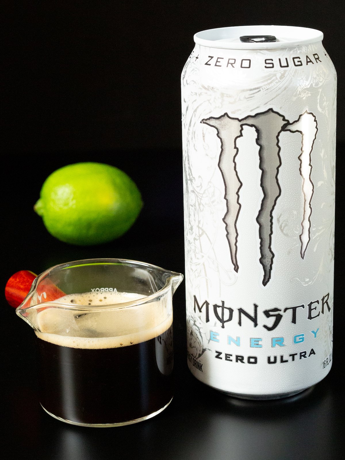 A small carafe of espresso, a white Monster energy drink, and a lime all on a black background.