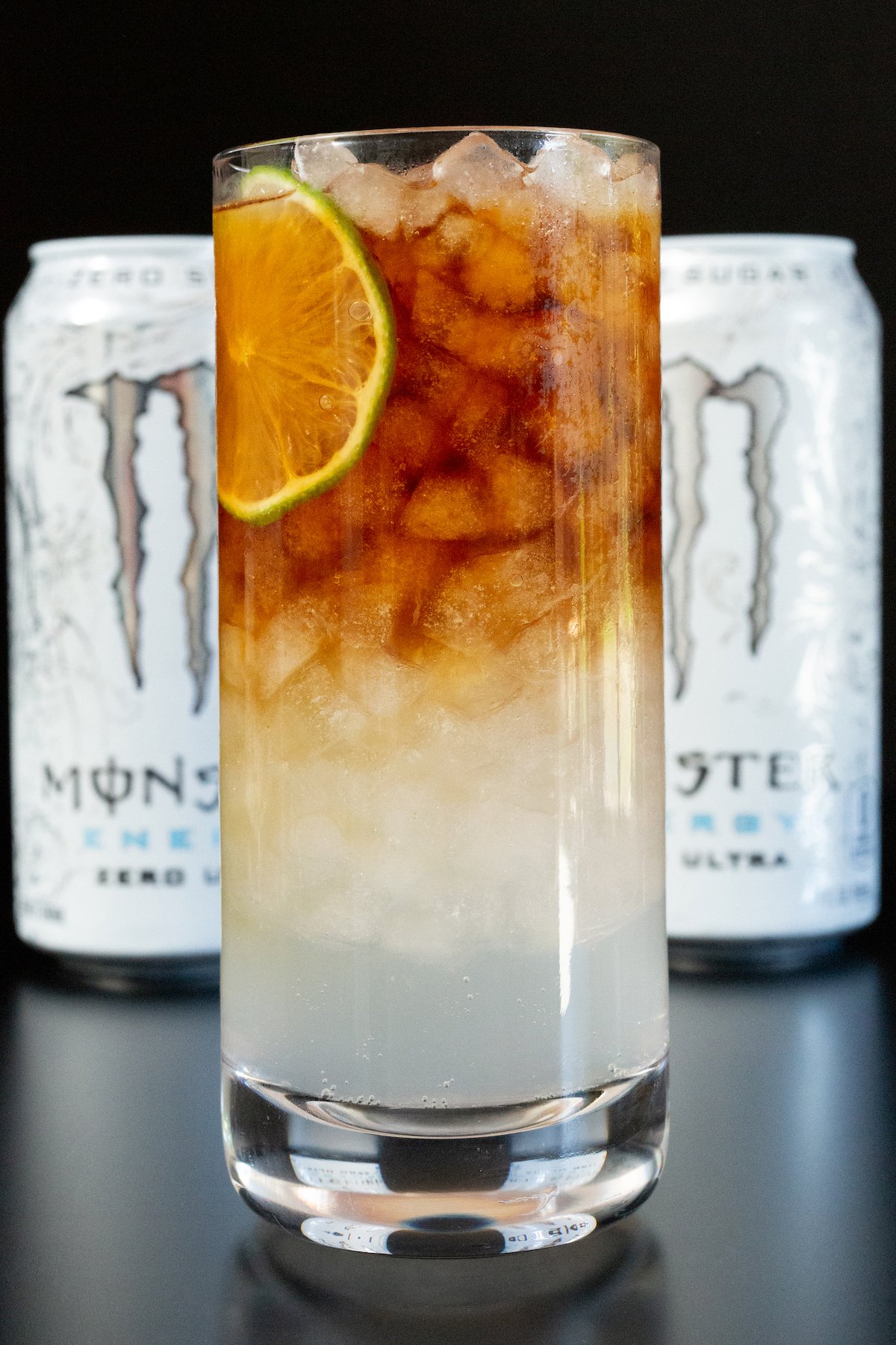 A highball glass filled half with white Monster and half with espresso, garnished with a lime wheel. Two cans of the energy drink are out of focus in the background.