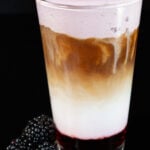 A pint glass is filled with a latte that has blackberry syrup at the bottom, milk in the middle, espresso, and a topping of blackberry cold foam. A pile of blackberries are out of focus next to the pint glass.