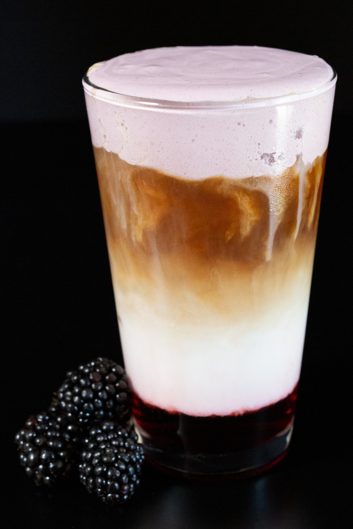A pint glass is filled with a latte that has blackberry syrup at the bottom, milk in the middle, espresso, and a topping of blackberry cold foam. A pile of blackberries are out of focus next to the pint glass.