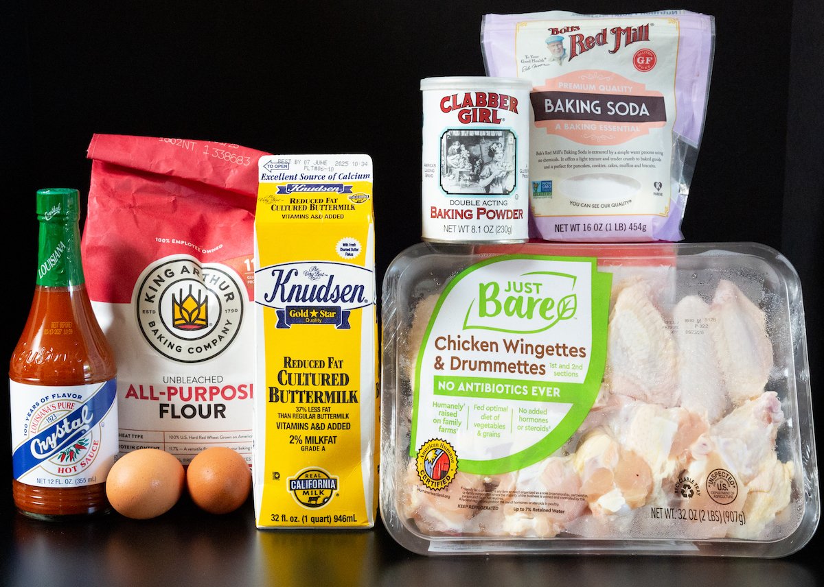 Raw chicken wings, a container of buttermilk, all-purpose flour, baking soda, baking powder, 2 eggs, and Crystal hot sauce all on a black background.