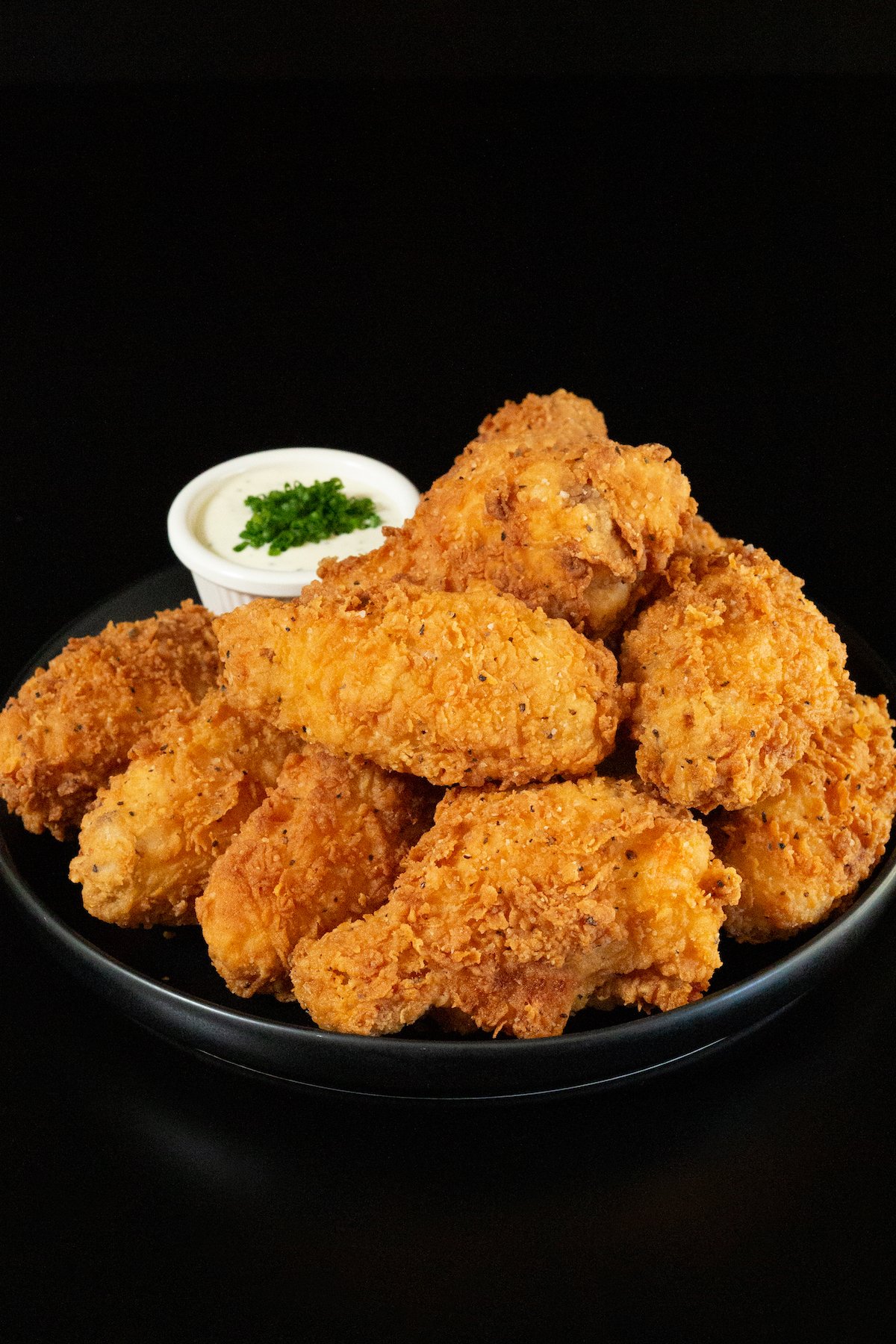 A small black plates has about a dozen breaded and fried chicken wings stacked on top of each other. A small side of ranch sauce topped with chives is out of focus in the background.