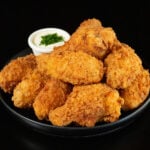 A small black plates has about a dozen breaded and fried chicken wings stacked on top of each other. A small side of ranch sauce topped with chives is out of focus in the background.