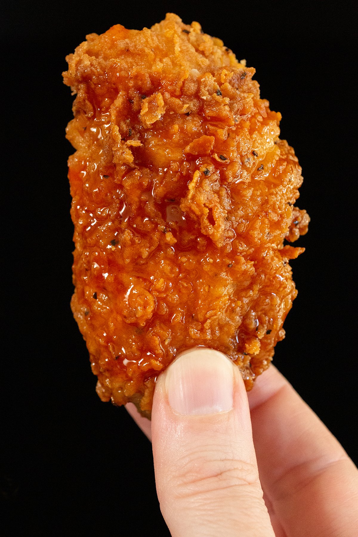 A hand holds up a single breaded chicken wings that's generously coated in hot sauce and honey.