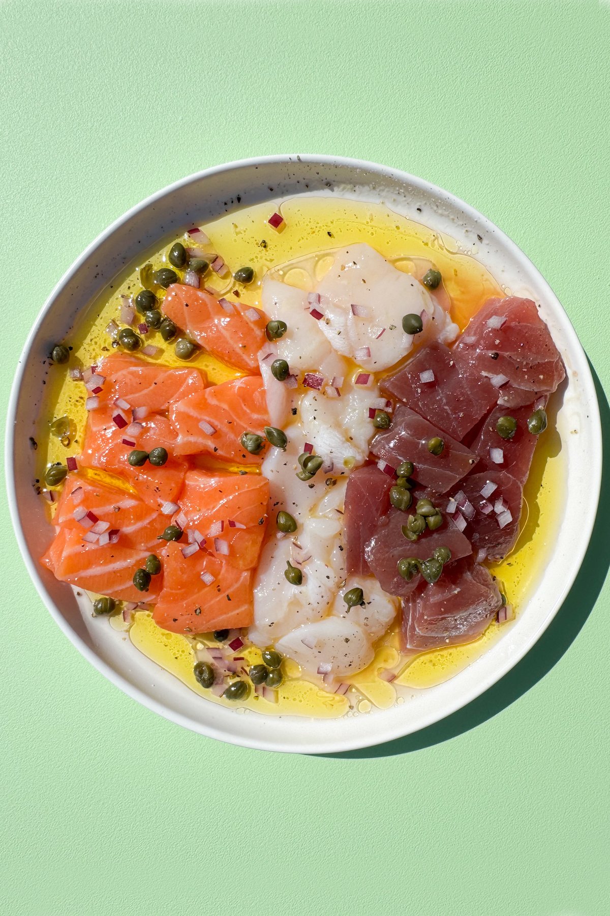 A small white plate with a row of bite-sized raw salmon pieces, raw tuna pieces, and slices of raw scallop surrounded by olive oil and lemon juice. Topped with diced red onion and capers.