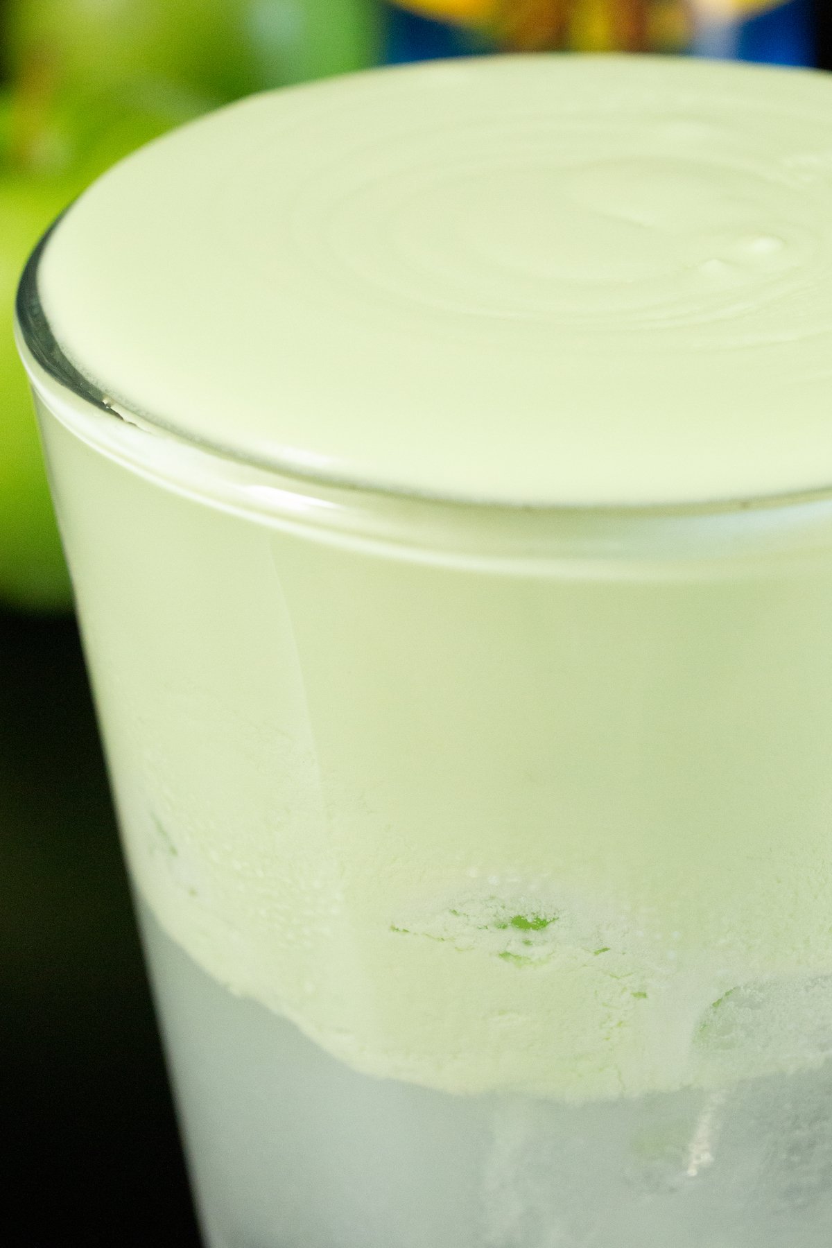 A pint glass is filled with a clear soda and generously topped with a thick and creamy green cold foam. Green apples are out of focus in the background.