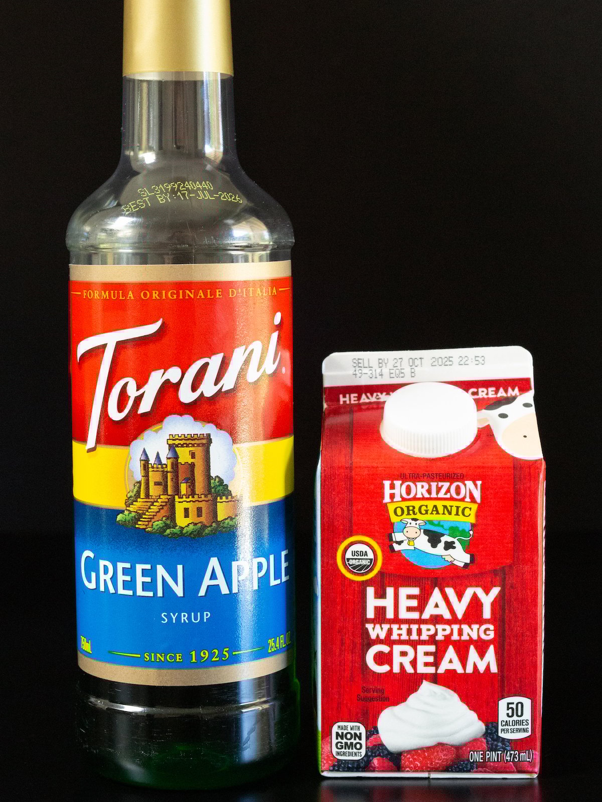 A bottle of Torani brand green apple syrup and a pint of organic heavy cream on a black background.