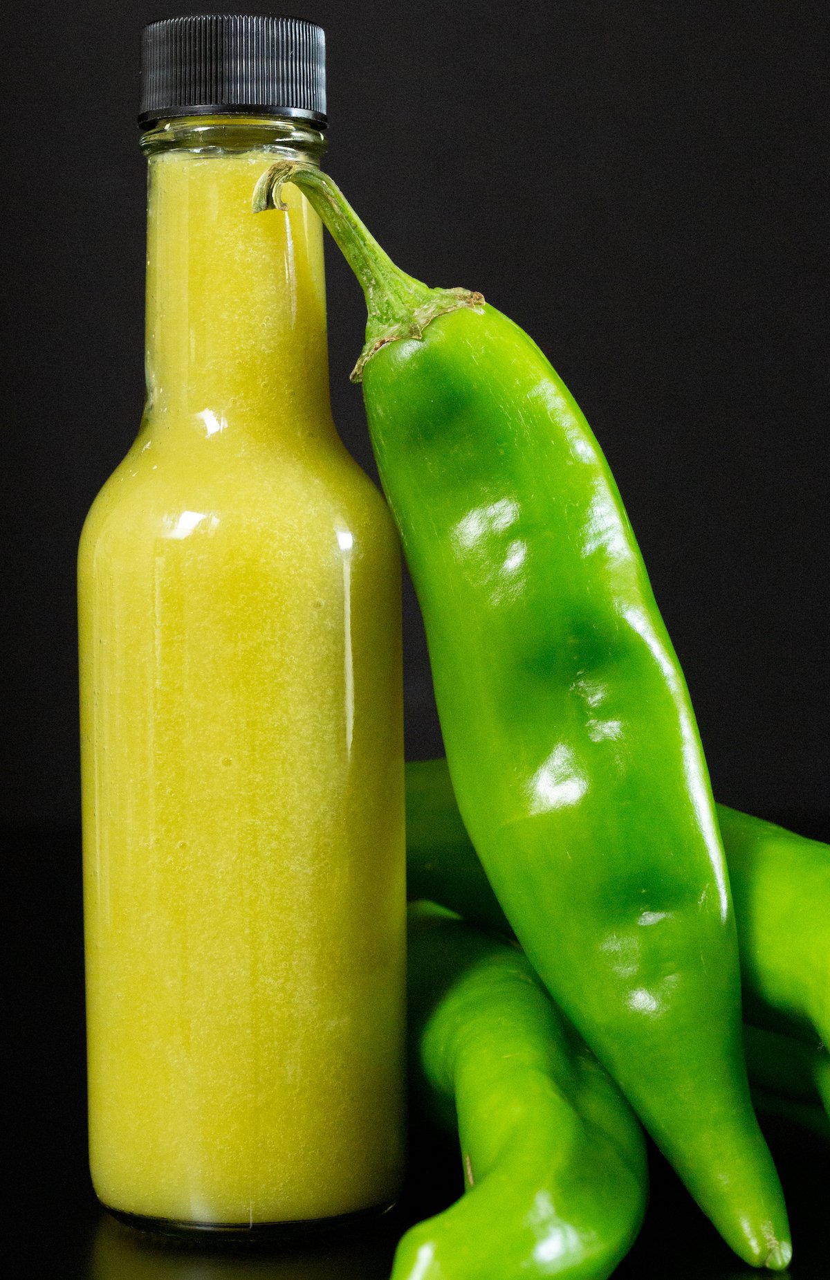 A 4 oz glass bottles is filled with a yellow-green hot sauce. Several fresh green hatch chiles are next to it.