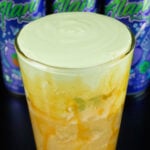 A pint glass is lined with caramel syrup and filled 2/3 of the way with brown soda, the top 1/3 is green apple cold foam. Three cans of Alani witches brew are out of focus in the background.