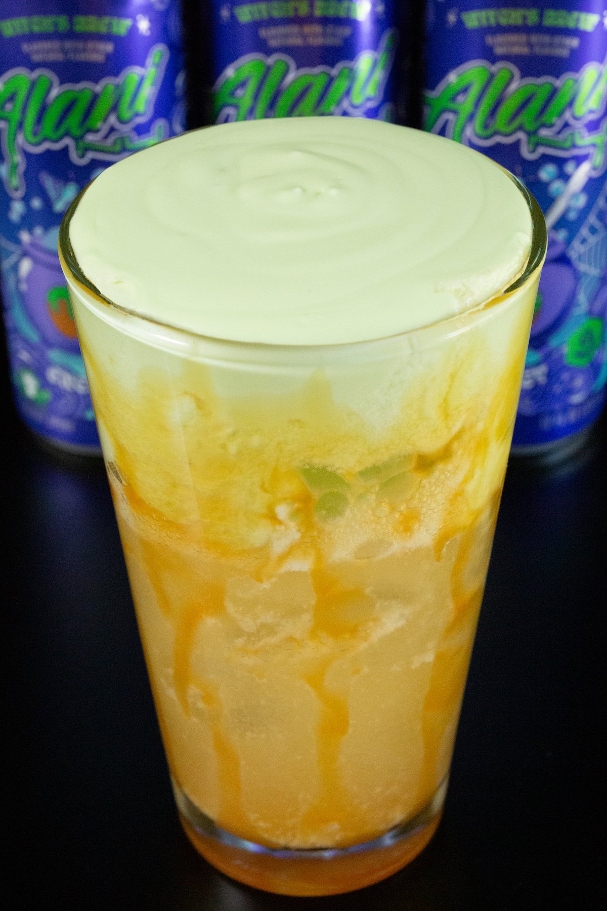 A pint glass is lined with caramel syrup and filled 2/3 of the way with brown soda, the top 1/3 is green apple cold foam. Three cans of Alani witches brew are out of focus in the background.