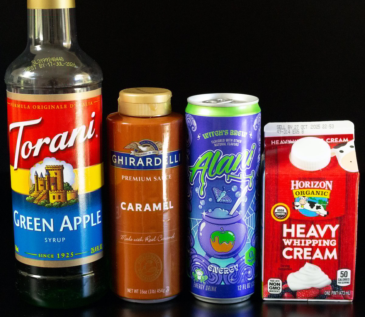 A bottle of Torani green apple syrup, Ghirardelli caramel sauce, a can of Alani Witch's Brew energy drink, and a pint of heavy cream.