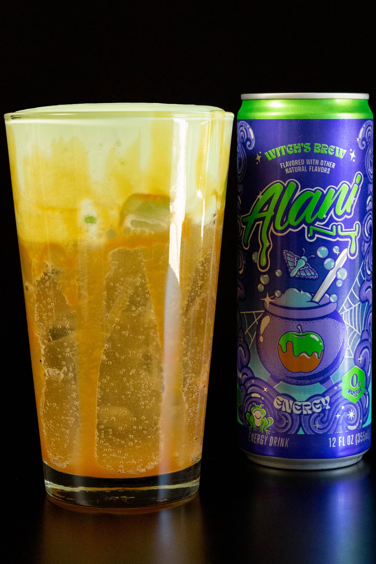 A pint glass is filled with a light brown energy drink 2/3 of the way. The top 1/3 is a green cold foam. Visible caramel sauce around the inside of the cup. A can of Alani witch's brew energy drink sits in the background.
