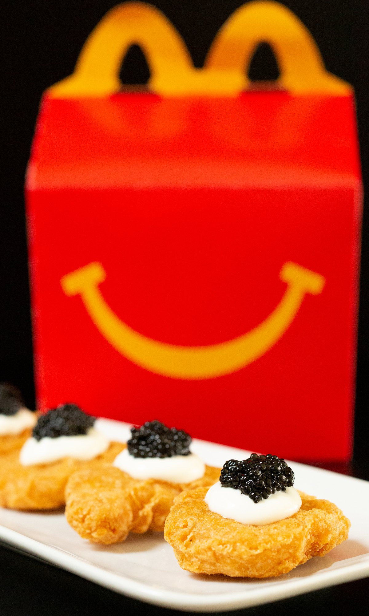 A small white plate filled with a row of Mcdonald's nuggets - each is topped with a dollop of creme fraiche and caviar. A happy meal box is out of focus in the background.