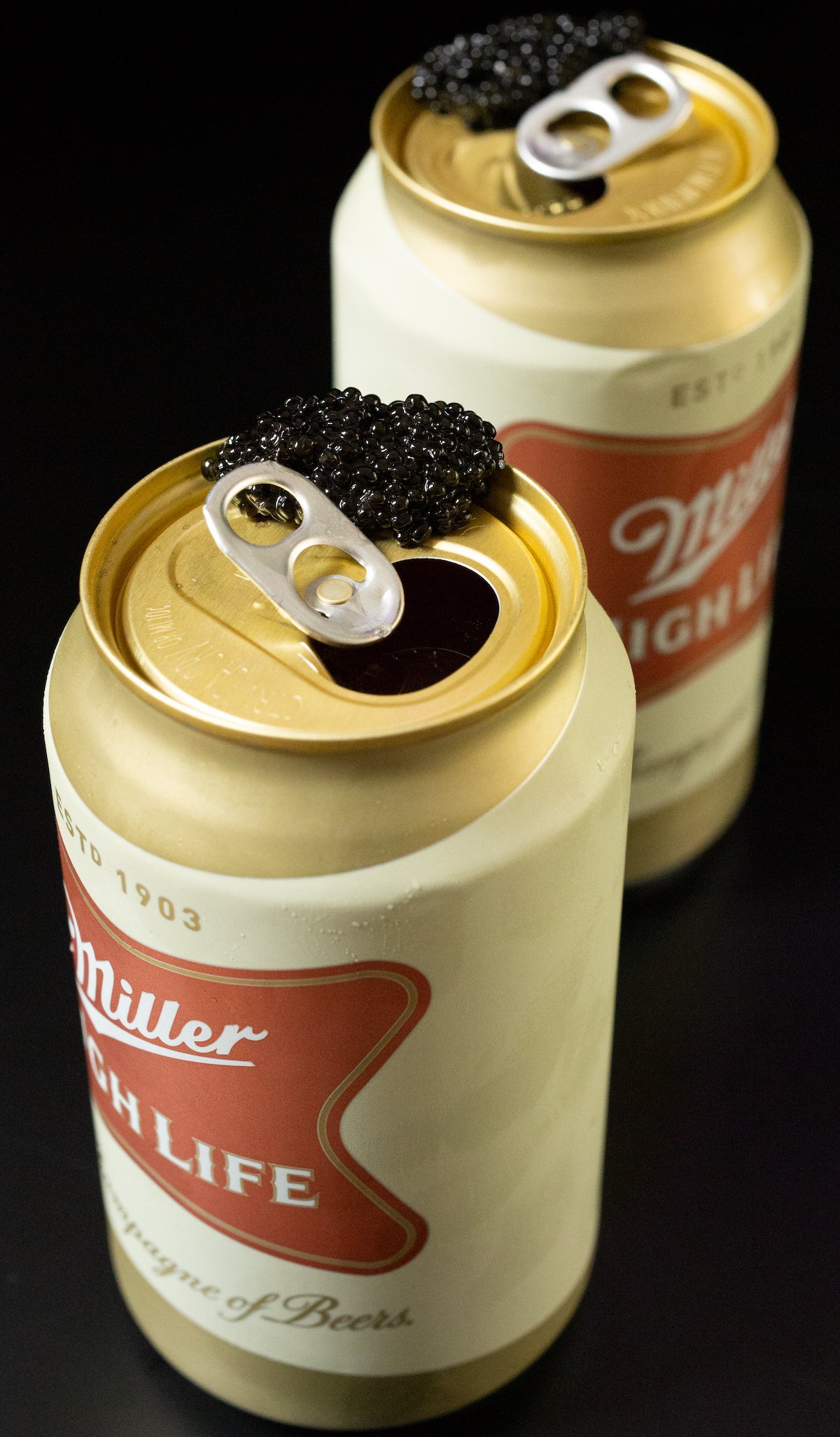 Two Miller High Life cans each with a generous bump of caviar on the rim.