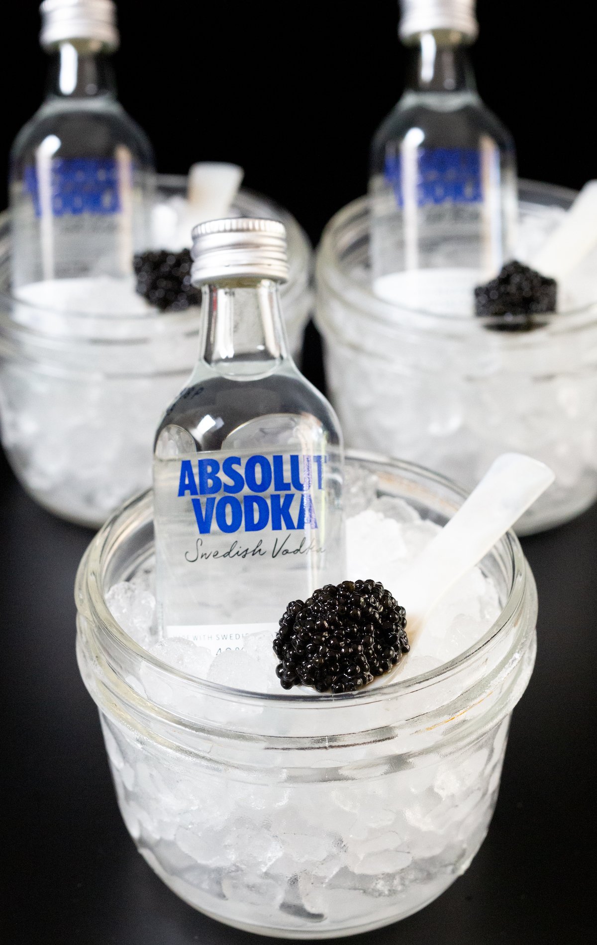 Three small glass jars filled with pebble ice each with a mini bar bottle of vodka and a pearl spoon with caviar.