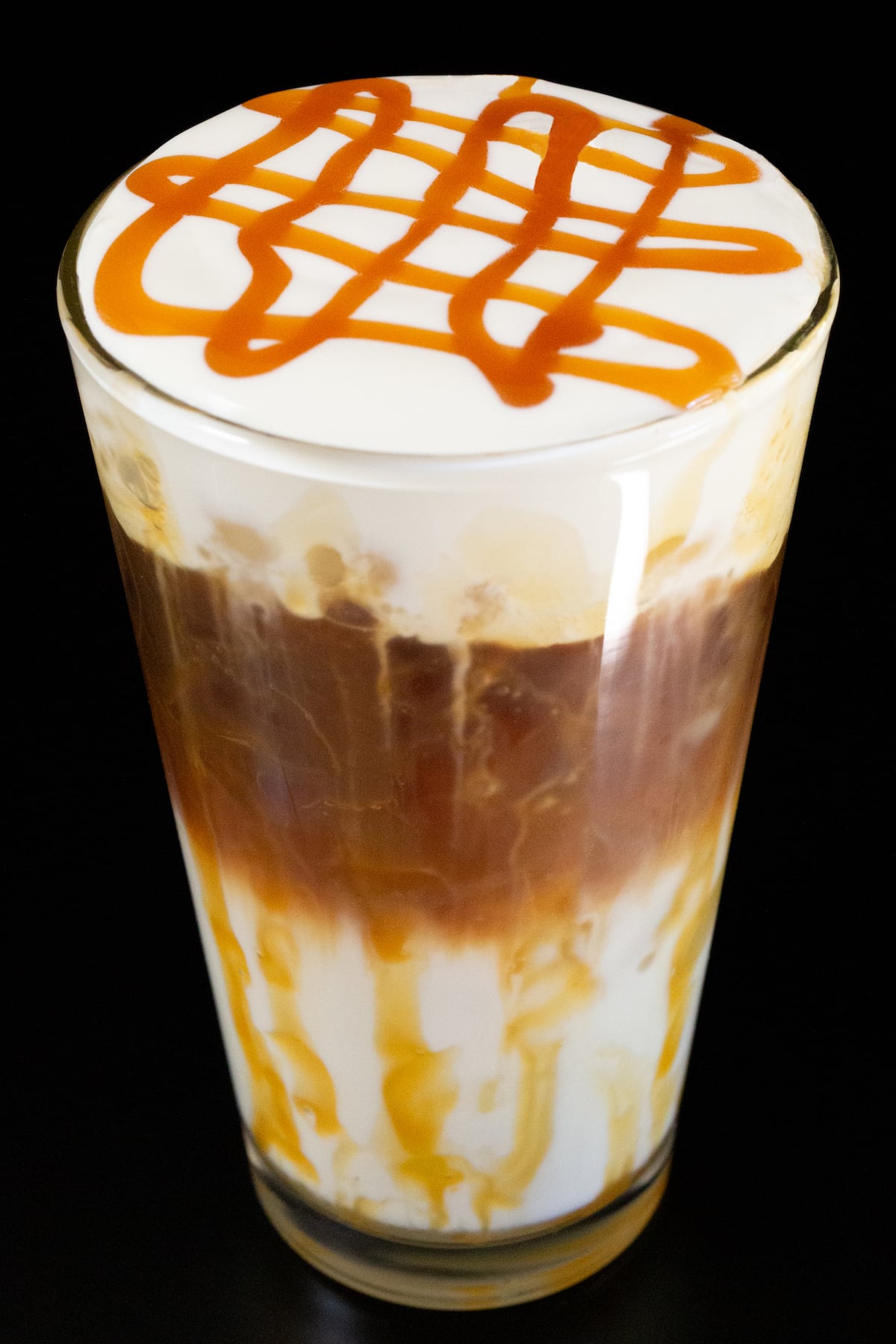 A pint glass has caramel drizzled all around the rim and is filled 1/3 of the way with milk, another 1/3 with espresso, then topped with a thick cold foam that has caramel drizzle on top.