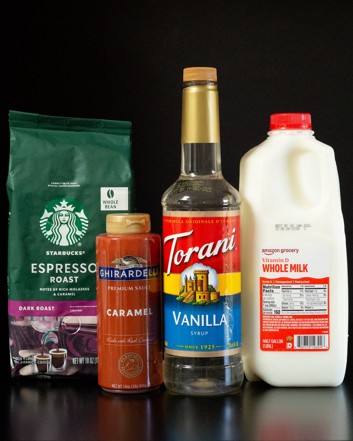 A bag of Starbucks espresso roast beans, a bottle of caramel sauce, a bottle of vanilla syrup, and a half gallon of whole milk on a black background.
