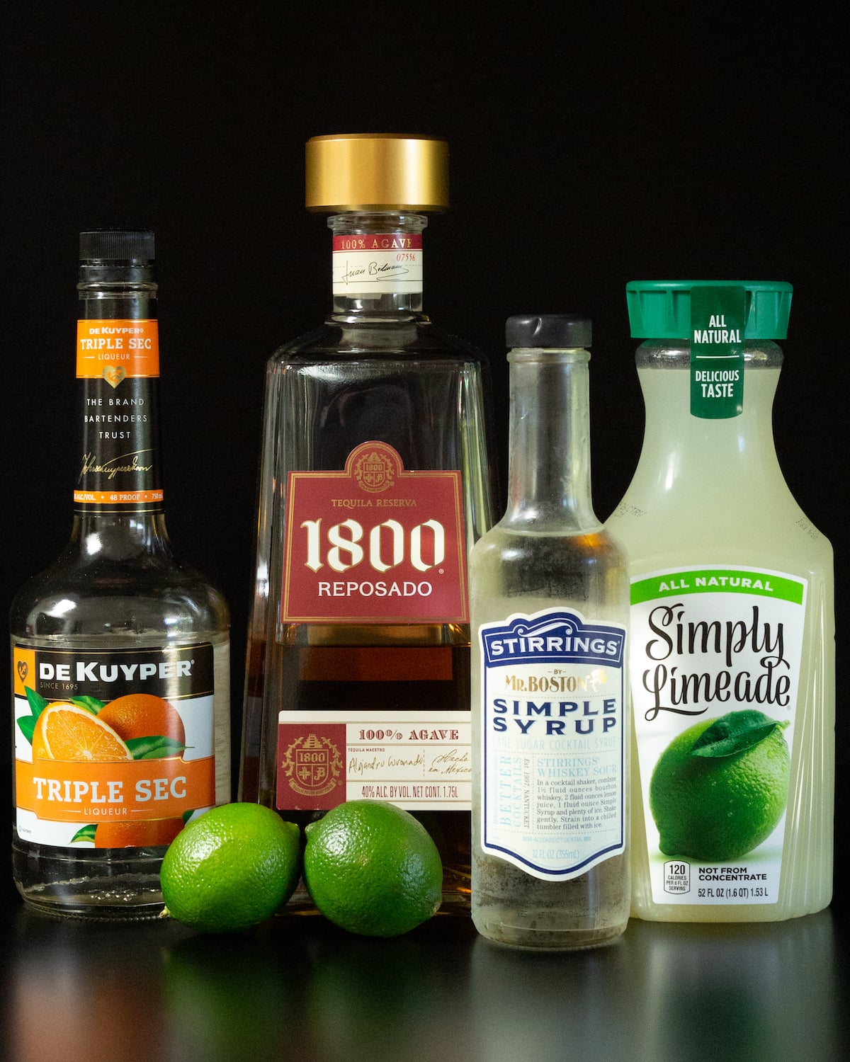 A bottle of of triple sec liqueur, 1800 tequila, simple syrup, and simply limeade next to two limes on a black background.