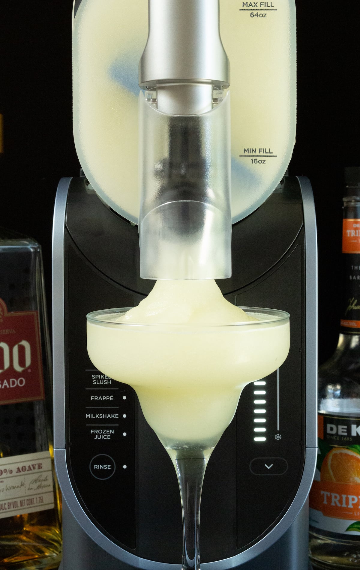 A Ninja Slushi machine has a tall margarita glass underneath its spout. It is being filled with a frozen margarita.